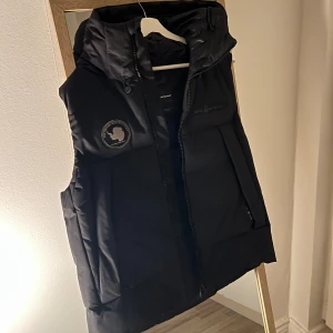 Svart dunväst från Sail Racing M - Svart dunväst från Sail Racing med huva och snyggt broderat märke på bröstet. Västen har dragkedja framtill och två stora fickor. Perfekt för kalla dagar och har en clean, sportig look. Materialet är polyester och dun, vilket gör den både varm och lätt. Skick: 9/10 Inga skador 