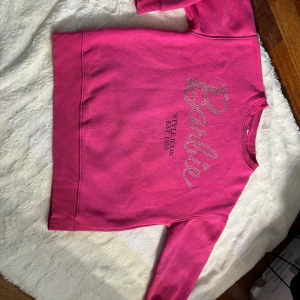 Rosa Barbie-tröja från Zara - Cool rosa tröja från Zara x Barbie med glittrande strassdetaljer som bildar Barbie-loggan och trycket 'STYLE ICON EST. 1959' på framsidan. Rund hals, ribbade muddar och långärmad. Perfekt för dig som älskar Barbie och vill sticka ut med färg.
