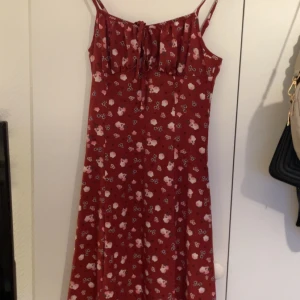 Röd blommig långklänning med smala axelband - Säljer en röd långklänning med blommigt mönster och smala axelband. Klänningen har en söt knytning framtill vid bysten och är ärmlös. Perfekt för sommarens varma dagar och har en flowy passform som ger en chill vibe.