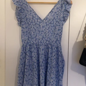 Blå blommig kortklänning med volang - Supersöt blå klänning med vitt blommönster och volangdetaljer längs axlarna. Djup v-ringning både fram och bak, samt en luftig kjol som ger en härlig sommarkänsla. Perfekt för dig som gillar romantiska och lekfulla plagg.