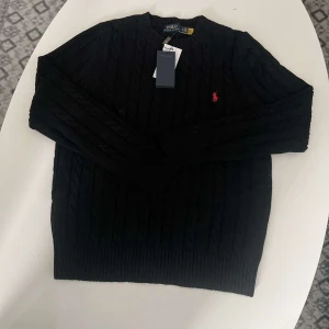 Svart kabelstickad tröja Polo Ralph Lauren - Svart kabelstickad tröja från Polo Ralph Lauren med klassisk röd broderad logga på bröstet. Tröjan har rund halsringning, ribbade muddar och är tillverkad i mjukt material som passar perfekt till lager-på-lager. Snygg och tidlös stil.
