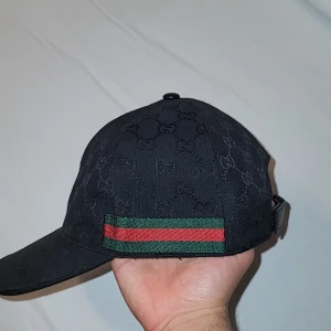 Gucci Keps  - Säljer en svart keps från Gucci med klassiskt GG-monogram över hela kepsen. Kepsen har justerbar rem bak i skinn och ikoniska gröna och röda ränder på sidan. Materialet är bomull och detaljer i skinn. Perfekt för dig som vill ha en lyxig och stilren accessoar.