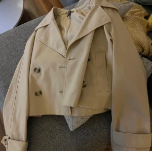 Beige croppad trenchcoat med knappar - Snygg croppad trenchcoat i beige med dubbelknäppning och bred krage. Jackan har långa ärmar med detaljer vid ärmslut och en rak passform. Perfekt för dig som vill ha en klassisk men trendig look.
