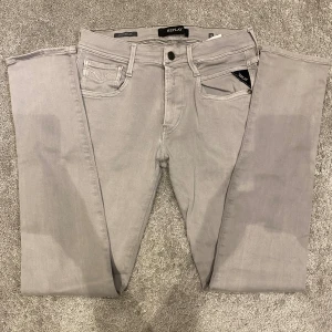 Replay Anbass Jeans - Säljer ett par ljusgrå Replay jeans i slim fit-modell. Jeansen har klassisk femficksdesign, dragkedja och bälteshällor. Snyggt broderad bakficka och mjukt stretchigt material som sitter skönt. Perfekta för dig som gillar stilrena och moderna jeans.