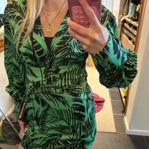 Grön mönstrad kortklänning Gina Tricot - Snygg grön kortklänning från Gina Tricot med svart palmbladsmönster. Klänningen har v-ringning, långa puffärmar och markerad midja. Tillverkad i ett lätt och luftigt material, perfekt för en trendig look.