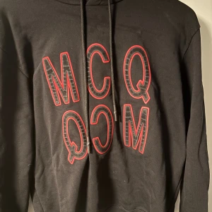 Svart hoodie från Alexander McQueen med MCQ röd logga - Svart hoodie från Alexander McQueen med stor, upp och nedvänd MCQ-logga i rött och svart på bröstet. Tröjan har huva med snörning och är tillverkad i mjuk bomull. Perfekt för dig som gillar streetwear och vill sticka ut med en fet logga.