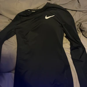Svart långärmad Nike Dri-Fit tröja - Svart långärmad Nike Dri-Fit tröja med tight passform. Tröjan har en vit Nike-logga på bröstet och är tillverkad i ett syntetmaterial som andas, perfekt för träning. Enkel och stilren design som sitter skönt mot kroppen.