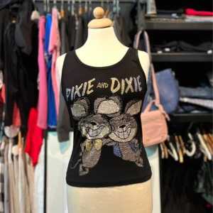 Svart linnetopp från Very Hot - Svart linnetopp från Very Hot med Pixie and Dixie-tryck fram och bak. Toppen har glittriga paljettdetaljer på texten och figurerna, samt breda axelband och rund halsringning. Perfekt för dig som gillar coola cartoon-motiv. Några stenar har ramlat av på texten bak (se bild 3)