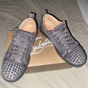 Christian Louboutin grå mocka sneakers - Säljer ett par grå sneakers från Christian Louboutin i mocka med ikoniska röda sulor. Skorna har snörning och coola nitar på tån för extra edge. Rund tå och platt sula, insida i beige skinn. Perfekt för dig som vill sticka ut med lyxig streetstil. Kan sänka priset vid snabb affär!