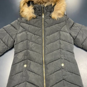 Svart pufferjacka med fuskpäls KappAhl - Svart pufferjacka från KappAhl med snyggt quiltat mönster och guldiga detaljer på dragkedja och fickor. Jackan har huva med fluffig fuskpäls och långa ärmar. Perfekt för kalla dagar och har en modern, slim passform.