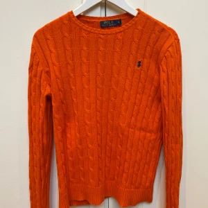 Orange kabelstickad tröja Polo Ralph Lauren - Snygg orange kabelstickad tröja från Polo Ralph Lauren i storlek S. Tröjan har rund hals, lång ärm och klassisk blå logga broderad på bröstet. Ribbstickade muddar vid ärmslut och nederkant. Perfekt för dig som gillar färg och stil.