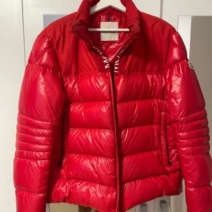 Röd Moncler pufferjacka med logga - Säljer snygg röd Moncler dunjacka. Jackan är kemtvättad och i mycket bra skick, med fin glans och fyllig dunvolym. Väldigt varm och bekväm. Observera: Den inre etiketten har lossnat men sitter ändå på jackan, när den har kemtvättas.  Se på sista bilden. Detta påverkar inte jackans utseende eller funktion. Storlek L 