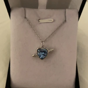 Halsband med blå hjärtberlock och pil silver - Supersött halsband med en silverfärgad kedja och ett hänge i form av ett blått hjärta genomborrat av en pil. Hjärtat är gnistrande och ger en romantisk vibe, perfekt för dig som gillar unika detaljer och vill sticka ut lite extra. Rostfri 