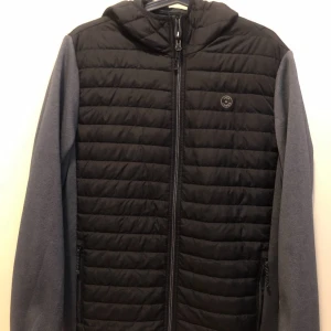 Svart och grå pufferjacka med huva - Snygg pufferjacka från Dope med svart quiltad front och rygg, grå ärmar och huva. Jackan har dragkedja framtill och två sidofickor med dragkedja. Perfekt för kyliga dagar när du vill hålla stilen enkel och clean.