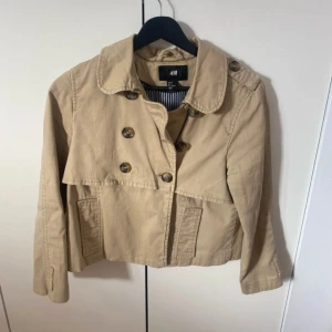 Beige kort trenchcoat från H&M - Kort trenchcoat i beige från H&M med dubbelknäppning, klassisk krage och snygga detaljer som axelklaffar och sprund bak. Jackan är tillverkad i bomull och har ett randigt innerfoder. Perfekt för dig som vill ha en stilren och tidlös look.
