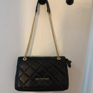 Svart quiltad axelväska från Valentino - Stilren svart axelväska från Valentino med quiltad design och guldfärgad kedja som axelrem. Väskan är tillverkad i skinn och har en elegant rektangulär form med logotypen i guld framtill. Perfekt för dig som gillar lyxiga detaljer och klassisk look.