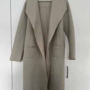Beige lång kappa med bälte - Snygg beige kappa med lång passform och bred krage. Jackan har två fickor framtill och ett matchande bälte som knyts i midjan. Materialet är mjukt och har en stilren look, perfekt för dig som gillar minimalistisk stil.