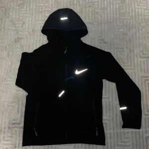 Svart vindjacka med huva. Nike 160-170cm - Svart vindjacka med huva och reflekterande detaljer, perfekt för träning utomhus. Jackan har dragkedja framtill, två sidofickor med dragkedja och en stor vit logga på bröstet. Materialet är lätt och syntetiskt, vilket gör den smidig att bära.