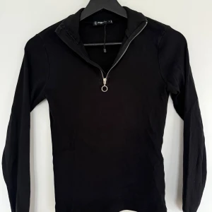 Svart ribbad långärmad half zip tröja FB Sister - Svart långärmad tröja från FB Sister i ribbad bomull med half zip-dragkedja och liten krage. Snygg och enkel modell som funkar till många olika stilar. Passformen är normal och materialet är mjukt och stretchigt. Aldrig använt