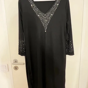 Svart långklänning med kristalldetajler - Svart långklänning i mjukt material med v-ringad hals och kristalldetaljer i ett V-mönster framtill samt på ärmarna. Klänningen har lång ärm och en rak, loose passform som ger en chill och stilren vibe. Perfekt för halloween outfit eller något annat