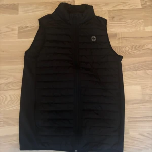 Svart dunväst från Jack & Jones - Svart dunväst från Jack & Jones med ståkrage och horisontella quiltade paneler. Västen har dragkedja framtill och en diskret logotyp på bröstet. Perfekt att slänga över en hoodie för extra värme under kyliga dagar.