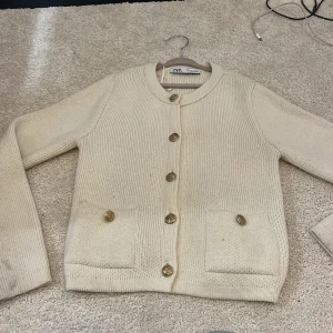 Beige stickad kofta från Zara - Snygg beige stickad kofta från Zara med rund hals och guldfärgade knappar framtill. Koftan har två fickor och långa ärmar, perfekt för lager-på-lager. Stilren och enkel design som funkar till många olika looks.