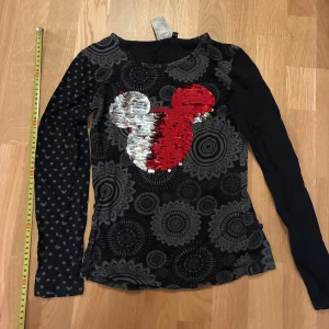 Desigual x Disney långärmad tröja - Unik långärmad tröja från Desigual x Disney med svart och grått mandalamönster, prickiga detaljer på ena ärmen och stor paljettdekor i rött och silver som formar Musse Pigg. Slim passform och mjukt bomullsmaterial. Från djurfritt rökfritt och prafymfritt hem! 