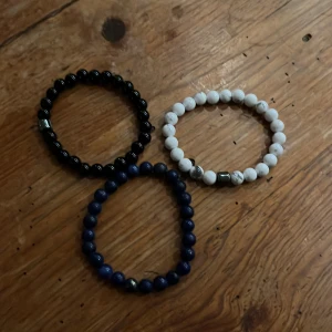 Set med tre pärlarmband i olika färger - Säljer ett set med tre elastiska armband av runda pärlor. Ett armband är svart med en metallisk detalj, ett är vitt med grå marmorering och ett är mörkblått. Perfekta för att mixa och matcha till din stil.