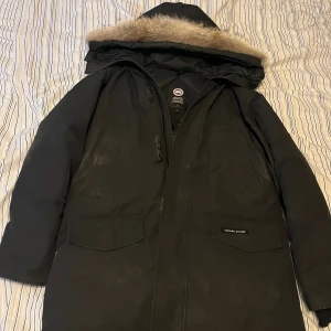 Svart dunjacka från Canada Goose - Säljer en svart dunjacka från Canada Goose med huva och beige pälsdetalj. Jackan har flera fickor, dragkedja och klassisk logga på ärmen. Perfekt för kalla vinterdagar och har en stilren look med lång passform.
