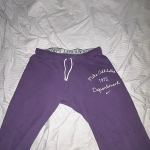 Lila mjukisbyxor från Nike - Säljer ett par lila mjukisbyxor från Nike med vit snörning i midjan och tryck på ena benet där det står 'Nike Athletic Department 1972'. Sköna och avslappnade med elastisk midja och mjukt material.