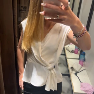 Vit omlottblus med knyt - Säljer en stilren vit omlottblus med knyt i midjan och v-ringning. Blusen är ärmlös och har en lätt, flowy känsla som ger en snygg siluett. Perfekt för dig som gillar en clean och fräsch look. Den har en liten fläck men den borde gå bort i tvätten