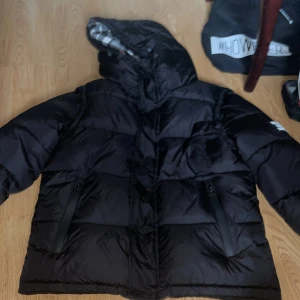  pufferjacka Burberry gamla  - Svart pufferjacka från Burberry med stor huva och klassiskt rutigt foder i grått, vitt och rött. Jackan har två fickor med dragkedja och en tydlig Burberry London England patch på ärmen. Storlek L men den är som en S plus den kan bli till en väst! Använt den några gånger men den är hel ny