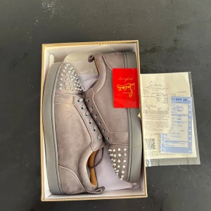 Gråa Christian Louboutin sneakers med nitar - Säljer ett par gråa sneakers från Christian Louboutin i mocka med ikoniska röda sulor och silverfärgade nitar på tån. Skorna har snörning och en riktigt clean look med lyxig känsla. Perfekta för dig som vill sticka ut med exklusiva detaljer.