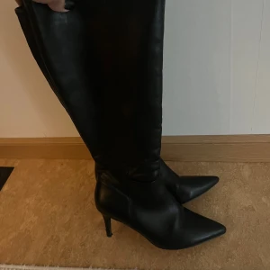 Svarta stövlar med spetsig tå och klack - Säljer ett par svarta höga stövlar i skinnimitation med spetsig tå och smal klack. Snyggt minimalistisk design som går upp till knäet och ger en riktigt clean look. Perfekta att styla med kjol eller jeans, otroligt bekväma, klacken har lite som behövs fixas, har haft de vid 2 tillfällen🖤