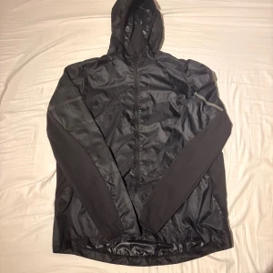 Svart vindjacka med huva från SOC XL - Svart vindjacka från SOC i storlek XL med huva och hel dragkedja. Jackan har tekniskt material som andas och blockerar vind, samt reflekterande detaljer på ärmar och rygg. Perfekt för blåsiga dagar och träning utomhus.