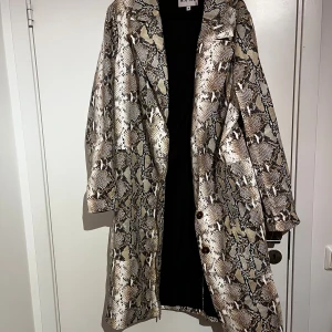 Ormmönstrad kappa från NA-KD - Snygg lång kappa från NA-KD med ormmönster i beige, brunt och svart. Rak passform, klassisk krage och knäppning framtill. 