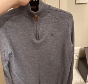 Grå/Blå merinoulltröja från Morris - Snygg långärmad tröja från Morris i 100% merinoull. Tröjan har en half zip med läderdetalj och en liten broderad logga på bröstet. Perfekt för dig som gillar stilrena och bekväma plagg med hög kvalitet.