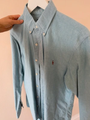 Ljusblå skjorta från Polo Ralph Lauren - Snygg ljusblå skjorta från Polo Ralph Lauren i slim fit. Klassisk button-down krage, broderad logga på bröstet och vita knappar. Tillverkad i mjuk bomull, perfekt för en clean och fräsch look. Passar dig som gillar stilrena plagg med premiumkänsla.