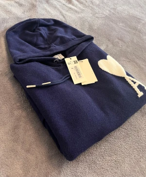 Ami Paris tröja  - Säljer en marinblå hoodie från Ami Paris med vit hjärtlogga och bokstaven A på bröstet. Tröjan har huva med snörning, ribbade muddar och magficka. Materialet är mjukt och stickat, perfekt för kyliga dagar. Stilren och modern design som sticker ut.