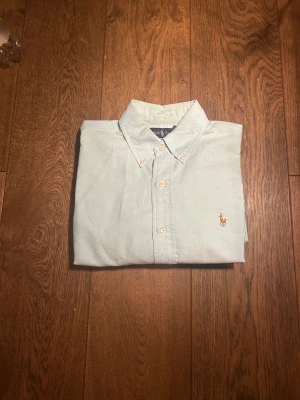 Ljusblå skjorta från Ralph Lauren - Storlek MEDIUM. Snygg ljusblå skjorta från Ralph Lauren med klassisk button-down krage och broderad logga på bröstet.Skjortan har knappar framtill och är tillverkad i mjuk bomull som känns skön mot huden. Perfekt för dig som gillar stilrena och tidlösa plagg.