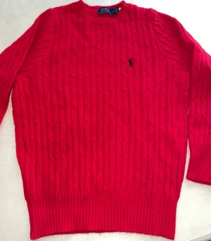 Röd stickad tröja Polo Ralph Lauren - Snygg röd stickad tröja från Polo Ralph Lauren med klassisk kabelstickad design och rund hals. Tröjan har lång ärm och ett broderat Polo-logo på bröstet. Perfekt för dig som gillar stilrena och tidlösa plagg med premiumkänsla.