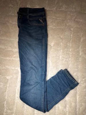 Replay Anbass blå jeans herr - Säljer ett par snygga Replay Anbass jeans i klassisk blå färg. Modellen har smal passform, fem fickor och coola detaljer som Replay-logga på fickan och bakfickor med V-söm. Jeansen är tillverkade i stretchigt bomullsmaterial för extra komfort.