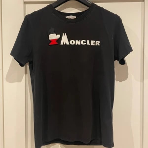 Svart Moncler t-shirt med logotyp - Svart t-shirt från Moncler med vit och röd broderad logotyp framtill. Klassisk rund hals och korta ärmar. Tillverkad i mjuk bomull med detaljer i viskos och silke för extra komfort. Snygg och stilren design som sticker ut med den ikoniska Moncler-loggan. Storleks lappen säger L men den sitter mer som en M