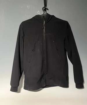 C.P. Company jacka - Svart softshelljacka från C.P. Company med ikoniska goggles i huvan. Jackan har dragkedja framtill, två snedställda fickor med dragkedja och ribbade muddar. Materialet är stretchigt och tillverkat av återvunnen nylon. Perfekt för dig som gillar techwear och streetstyle.