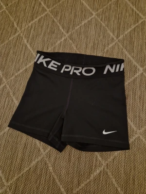 Nike Pro svarta träningsshorts - Svarta Nike Pro shorts med bred elastisk midja och vit logga framtill. Sitter tight och är perfekta för träning. Materialet är syntet och designen är enkel med diskreta sömmar. Midjan är hög och resåren har Nike Pro-tryck i vitt.
