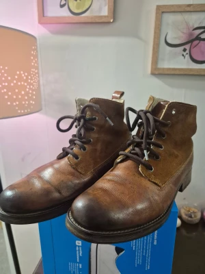 Roots Kängor Brun Storlek 42 i skinn och mocka - Säljer ett par bruna kängor med snörning och rund tå. Ovandelen är tillverkad i både skinn och mocka, vilket ger en schysst kontrast. Sulan är platt och robust, perfekt för dig som gillar klassisk stil med lite edge. Insidan är ljus och mjuk för extra komfort.