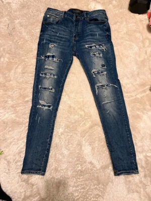 Amiri jeans - Säljer ett par blå skinny jeans från AMIRI med coola slitningar och patchade detaljer i rutigt tyg på framsidan. Jeansen har klassisk femficksdesign, normal passform och är tillverkade i stretchig bomull för skön känsla. Perfekt för dig som gillar en edgy streetstil.