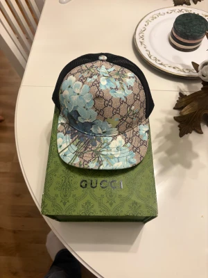 Gucci blommig keps med mesh - Snygg keps från Gucci med beige och brun bas, blå blommor och klassiskt GG-mönster. Framsidan är i canvas med blommigt tryck, baksidan i svart mesh för extra ventilation. Justerbar passform och märkning på insidan.