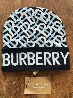 Burberry mönstrad mössa i svart och blått - Snygg mössa från Burberry med grafiskt mönster i svart, vitt och ljusblått. Stor logotyp framtill och uppvikt kant. Stickad i mjukt material som håller dig varm och ger en cool look. Perfekt för dig som vill ha något extra till vintergarderoben.