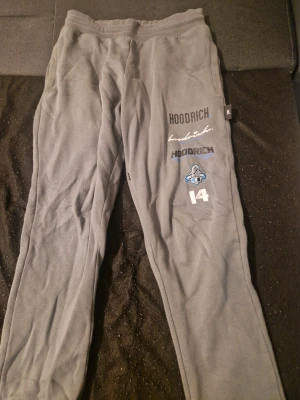 Grå Hoodrich mjukisbyxor med tryck - Säljer ett par grå mjukisbyxor från Hoodrich med flera tryck och broderade detaljer på framsidan och baksidan, bland annat 'Hoodrich', '14' och 'Hood Series'. Byxorna har elastisk midja och är gjorda i mjukt bomullsmaterial. Perfekt för chill och streetwear. (Har ett litet hål på knät)